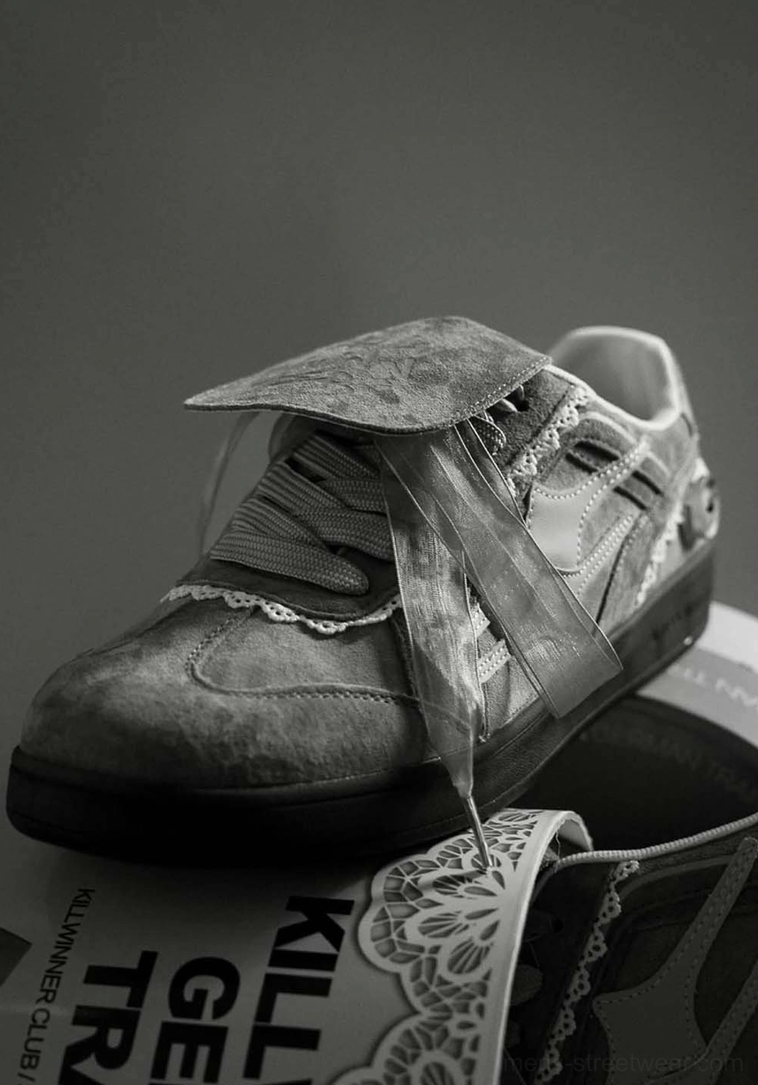 Close-up of grey lace-wrapped retro trainer sneaker showing textured upper, graphic tongue and decorative strap detail