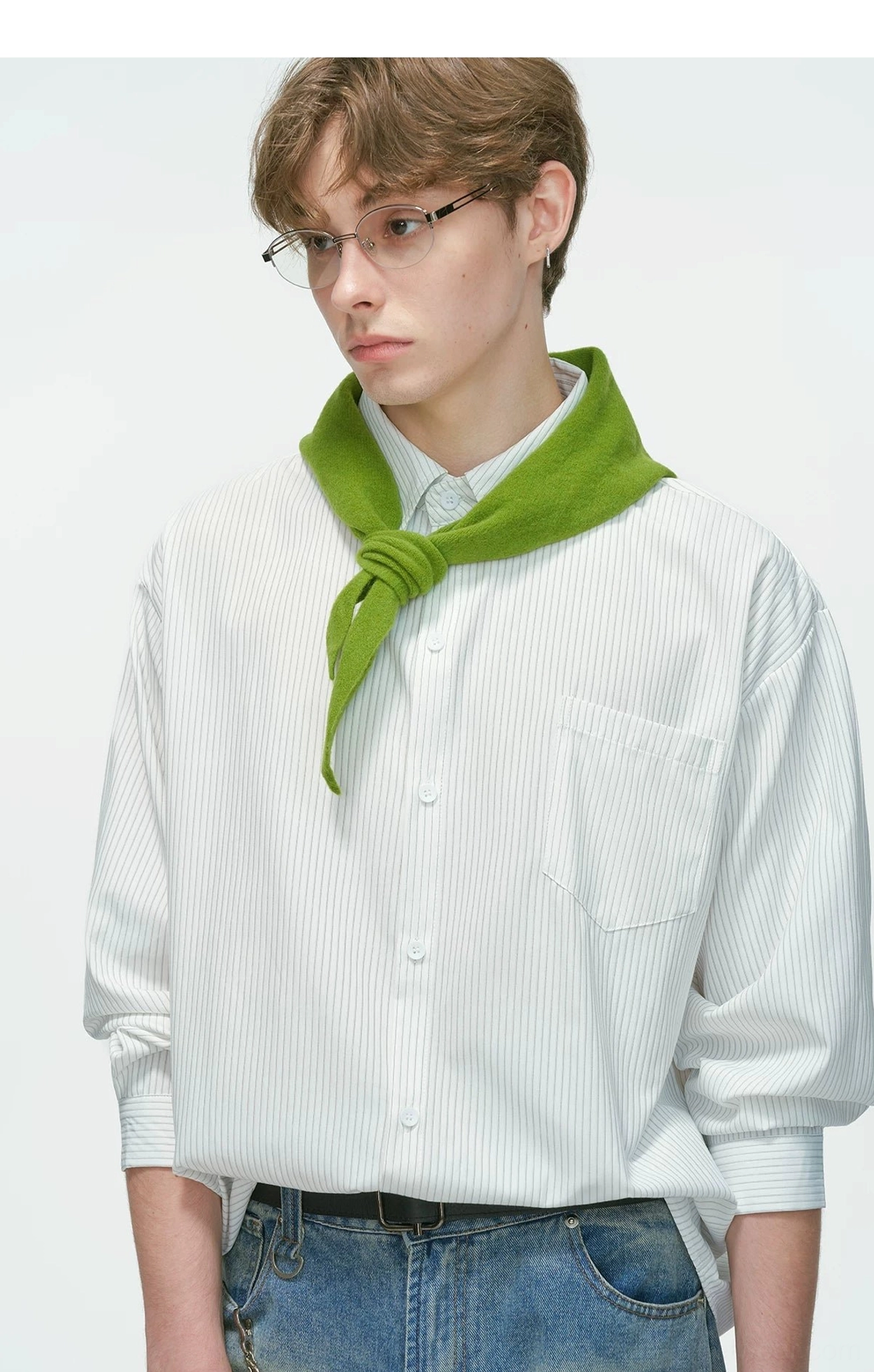 Close-up of a male model wearing a grey thin vertical stripe relaxed overshirt with pointed collar, blue jeans, and green neck scarf