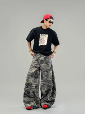 Full body side view of an oversized black Happiness Plan graphic T-shirt styled with camouflage wide leg pants red sneakers and a backward baseball cap