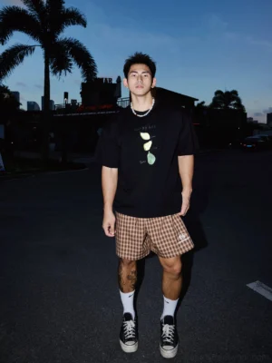 Front view of a black Happy Day citrus graphic T-shirt styled with checkered shorts, socks, and sneakers in a palm-lined streetwear setting