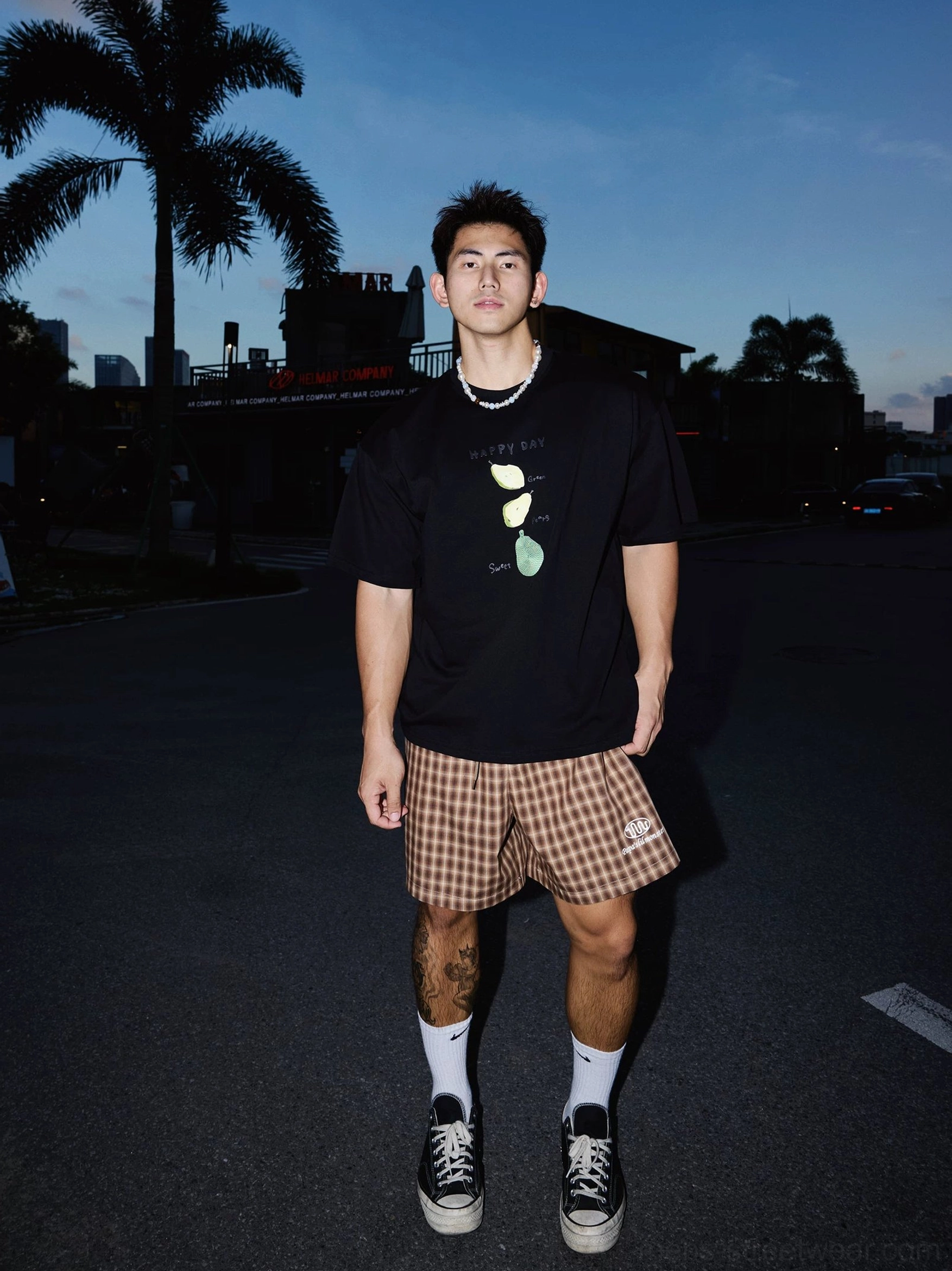Front view of a black Happy Day citrus graphic T-shirt styled with checkered shorts, socks, and sneakers in a palm-lined streetwear setting