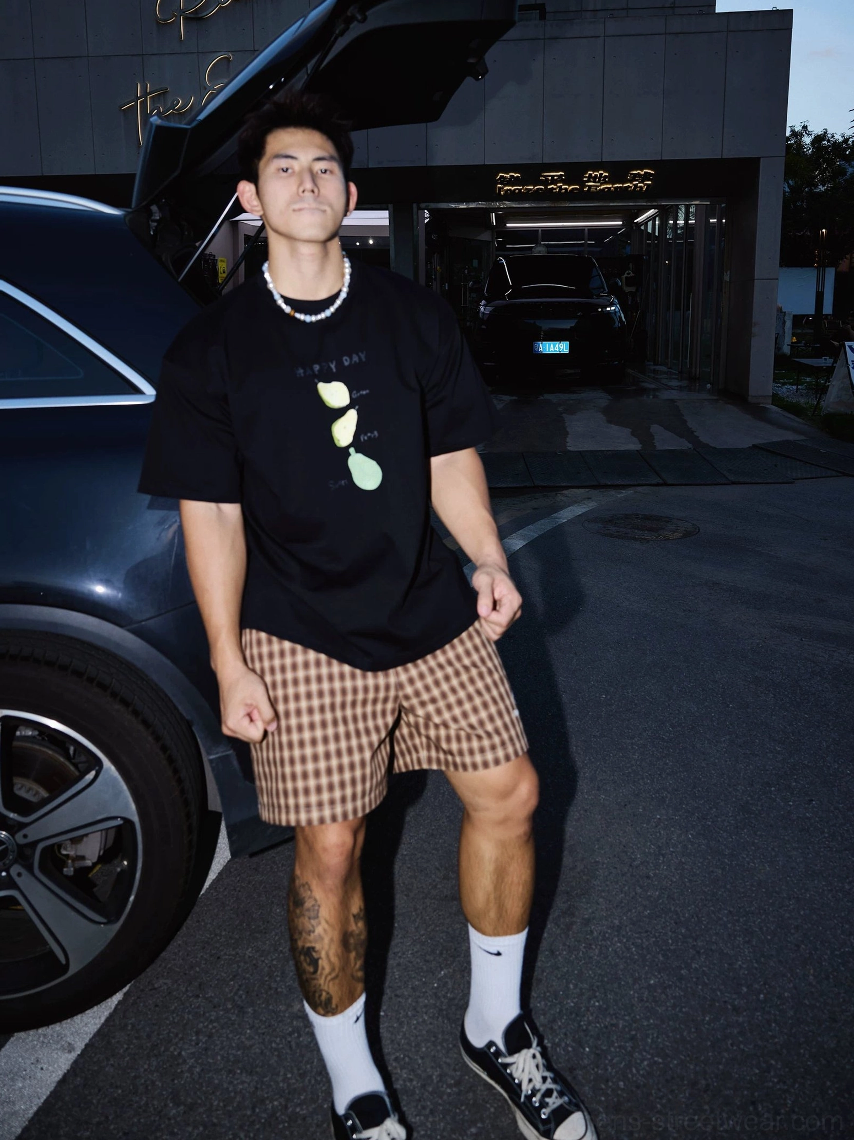 Three-quarter view of a black citrus graphic streetwear T-shirt with checkered shorts, canvas shoes, and pearl necklace styling