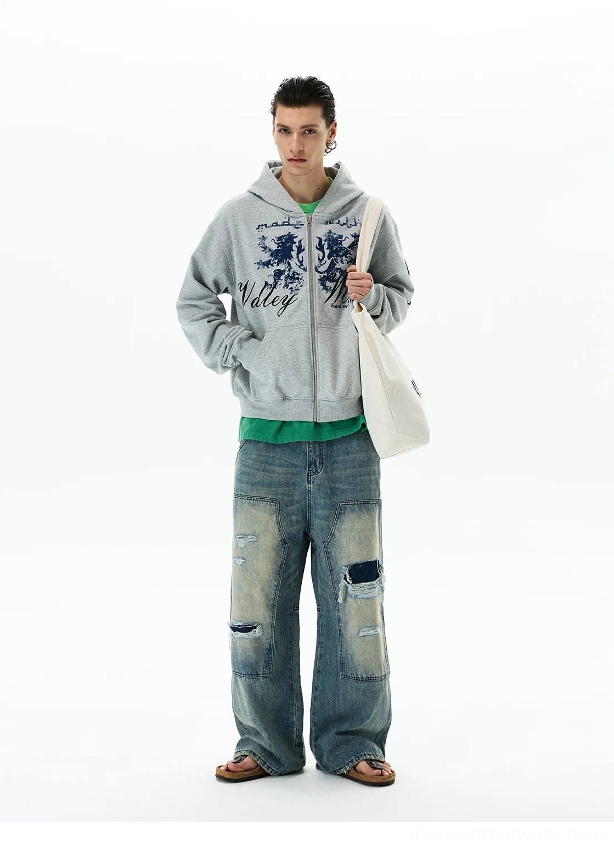 Front three-quarter view of a heather grey vintage crest zip hoodie styled with a green tee, ripped baggy jeans, and a white canvas tote