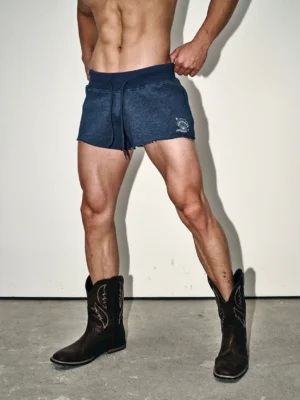 Front close-up of dark indigo raw hem cutoff sweat shorts with elastic drawstring waist and small white leg graphic styled with cowboy boots