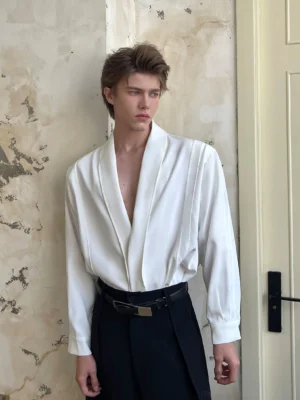 Men's ivory deep V drape shirt styled with black belted wide-leg pants in a retro indoor setting