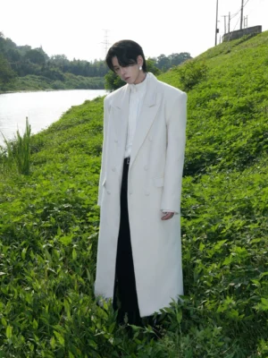 Men's ivory double-breasted wool blend long overcoat shown in left side view outdoors on a riverside grassy slope
