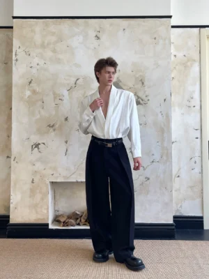 Full-body male model wearing an ivory satin drape shirt with black high-waisted trousers and leather belt