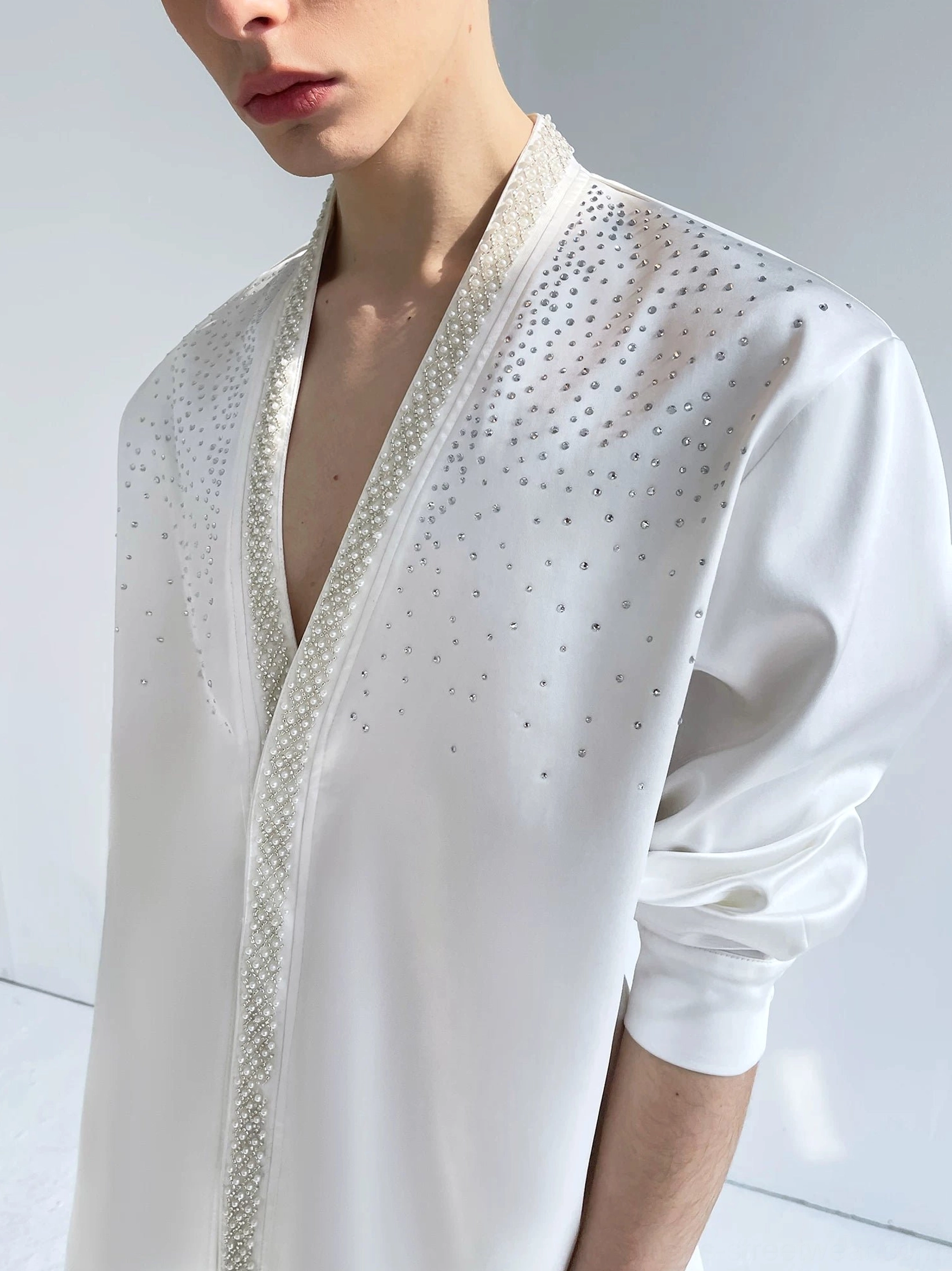 Close-up of an ivory satin men's shirt showing the pearl-style beaded front trim and scattered crystal rhinestone details across the chest and shoulder
