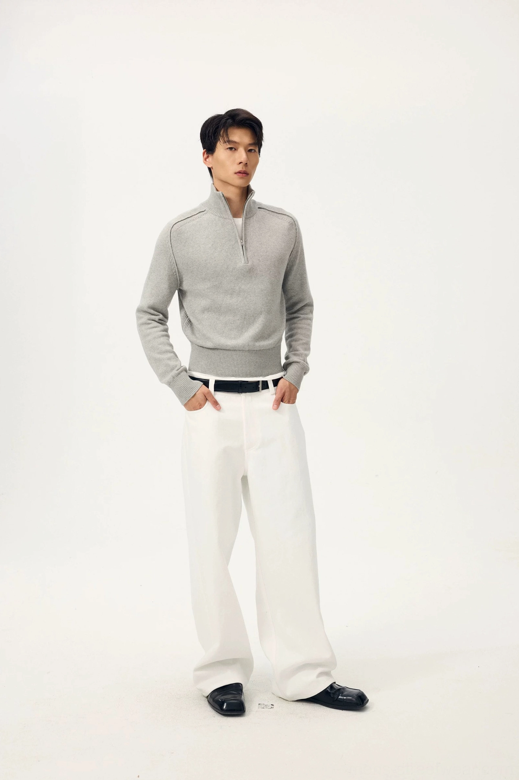 Male model wearing ivory white wide-leg denim trousers with a grey half-zip knit sweater, black leather belt, and loafers in a minimalist studio