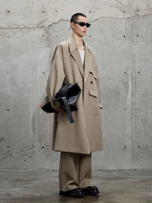 Khaki long trench coat shown from the left side with flap chest pockets, tailored trousers, sunglasses, black leather clutch, and loafers