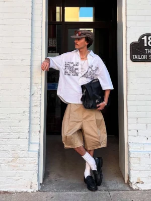 Male model wearing khaki loose-fit Bermuda shorts with printed shirt dress socks loafers and crossbody bag