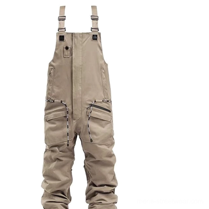 Khaki unisex utility bib snow pants displayed from the front with adjustable suspender straps, zippered pockets, relaxed snow-ready fit, and technical outdoor shell