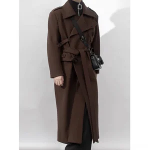 Male model wearing a dark brown oversized belted trench coat with black clothing, leather boots, and a crossbody bag in a white studio