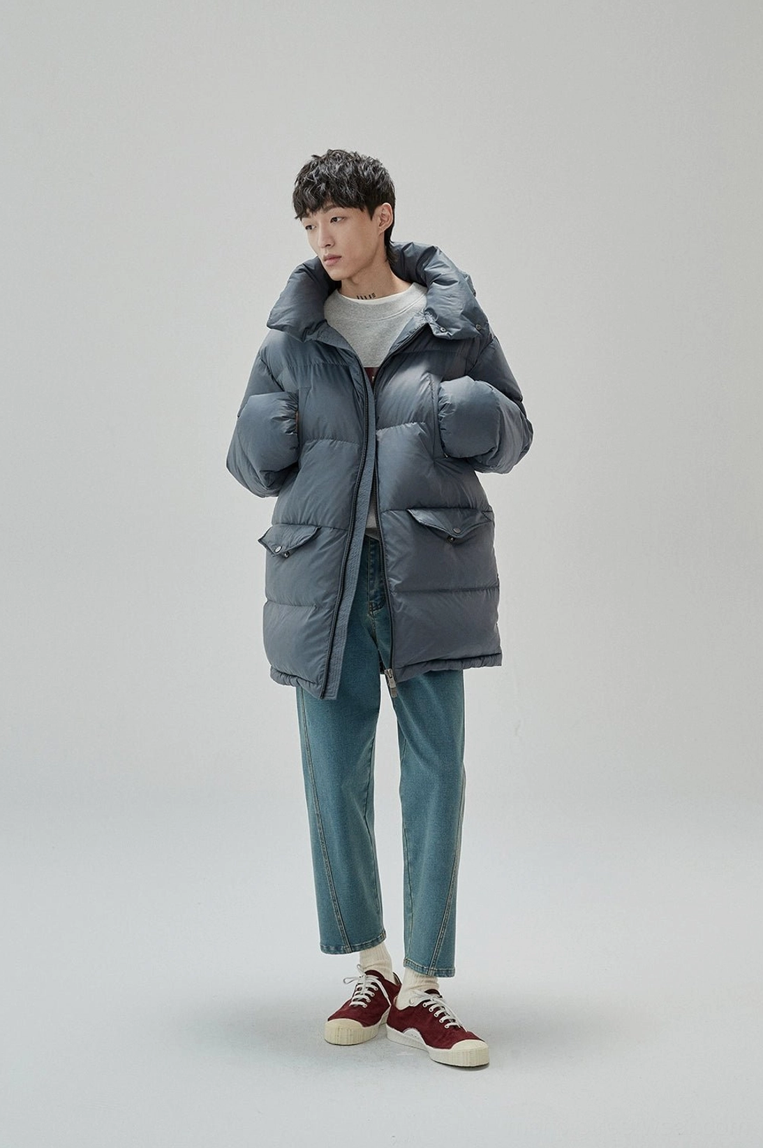 Side view of layered gray UNIFORM sweatshirt outfit with oversized down jacket blue jeans and burgundy sneakers