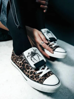 Close-up view of leopard print canvas streetwear sneakers being adjusted, showing the black spider embroidered tongue strap and lace-up closure