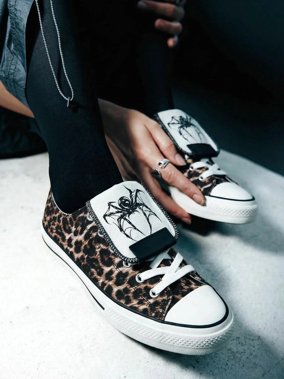 Close-up view of leopard print canvas streetwear sneakers being adjusted, showing the black spider embroidered tongue strap and lace-up closure