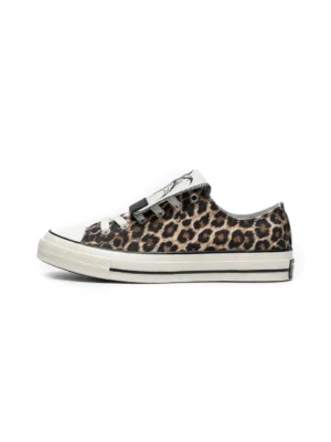 Side profile of leopard print low-top canvas sneakers with white rubber toe cap, flat sole, detachable spider embroidered tongue, and contrast laces