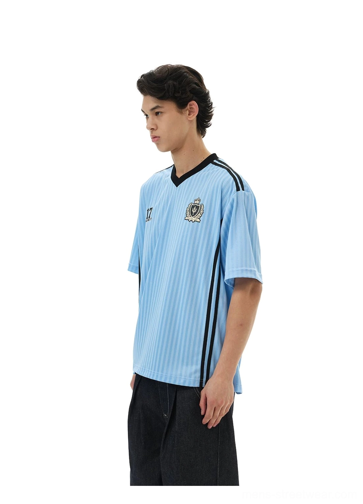 Light blue crested soccer jersey tee shown from the front side with black V-neck trim, shoulder stripes, and relaxed streetwear fit