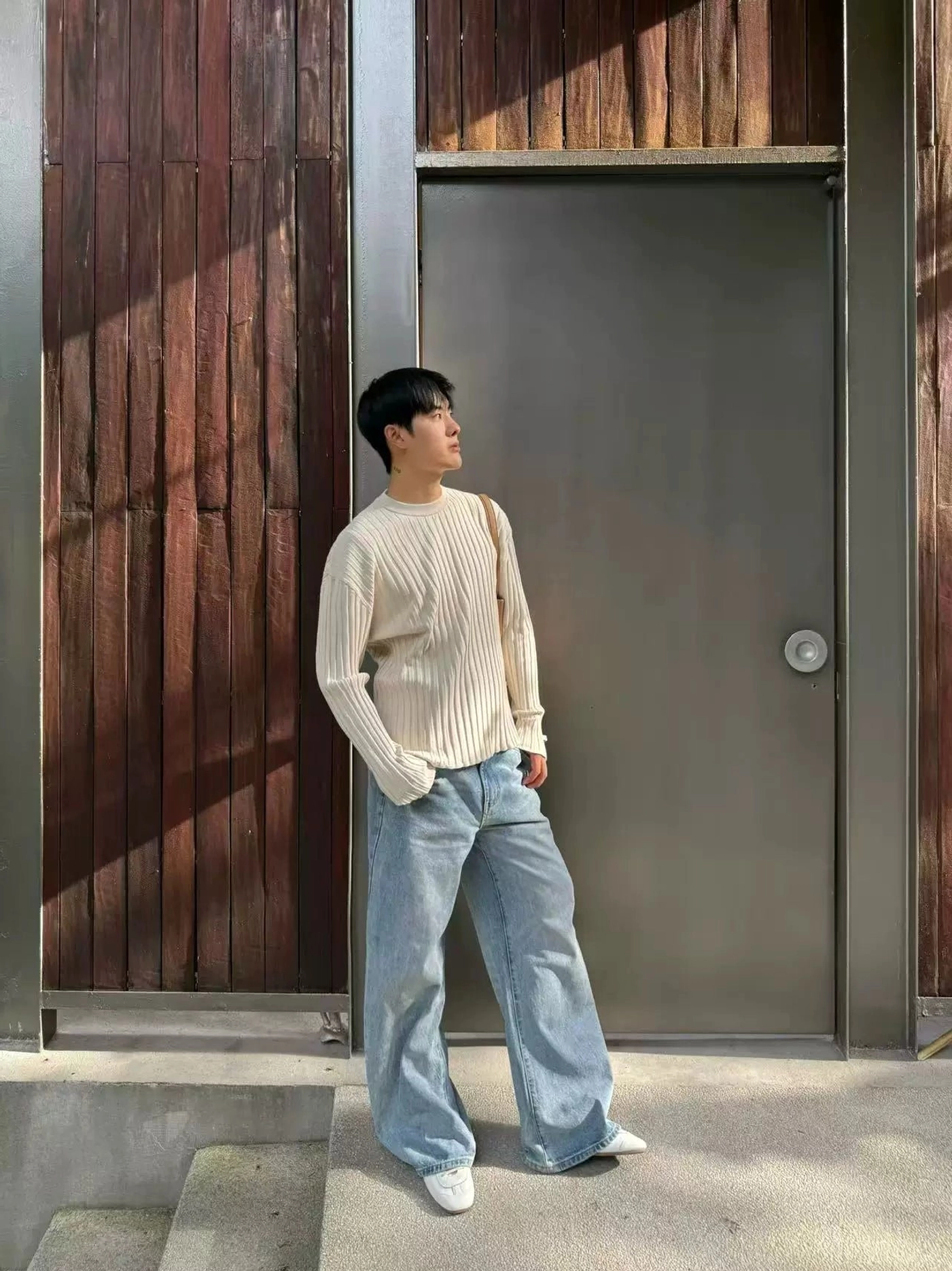 Side profile of light blue five-pocket wide leg denim jeans worn with a ribbed crewneck sweater at an outdoor residential entrance