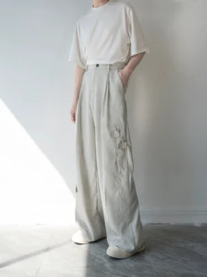 Front view of high-waisted light gray wide-leg tailored trousers with deep pleats, button closure, side pockets, and floral embroidery