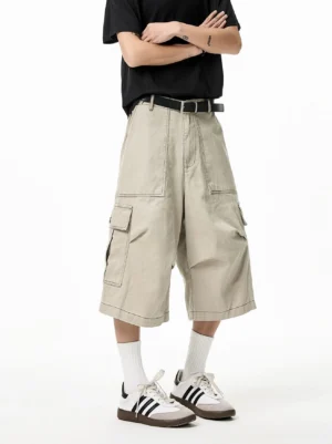 Light khaki oversized cargo Bermuda shorts shown from the front with wide relaxed legs, utility flap pockets, clean canvas fabric, black T-shirt, white socks, and stripe sneakers