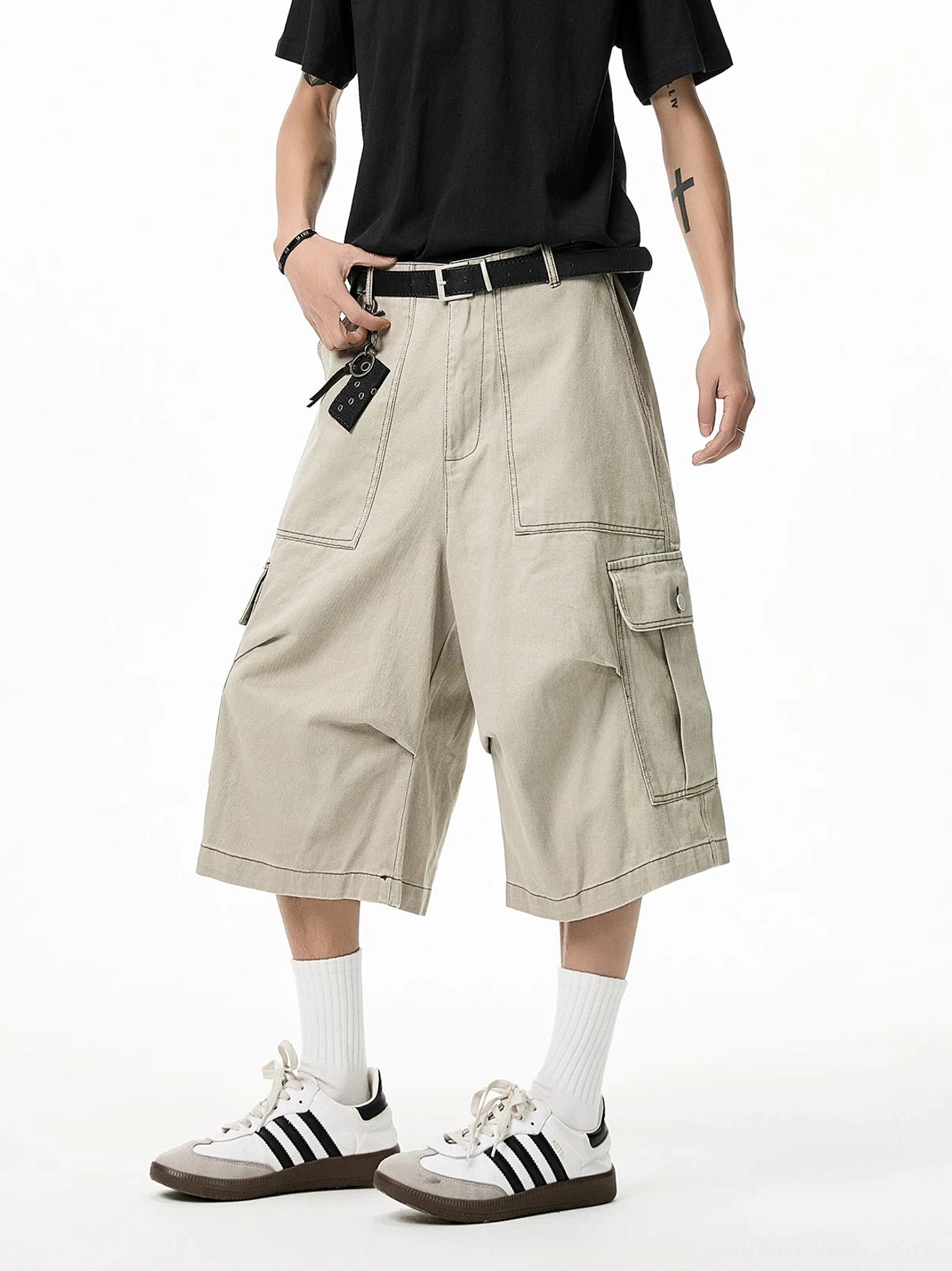 Light khaki wide-leg cargo Bermuda shorts shown in a three-quarter view with multiple flap pockets, exposed topstitching, black T-shirt, belt accessory, and stripe sneakers