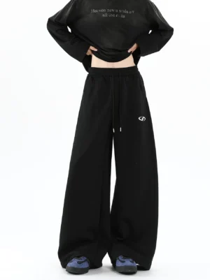 Model wearing black logo wide-leg sweatpants with adjustable drawstring waist, cropped graphic long-sleeve top, and casual sneakers in a Korean streetwear outfit