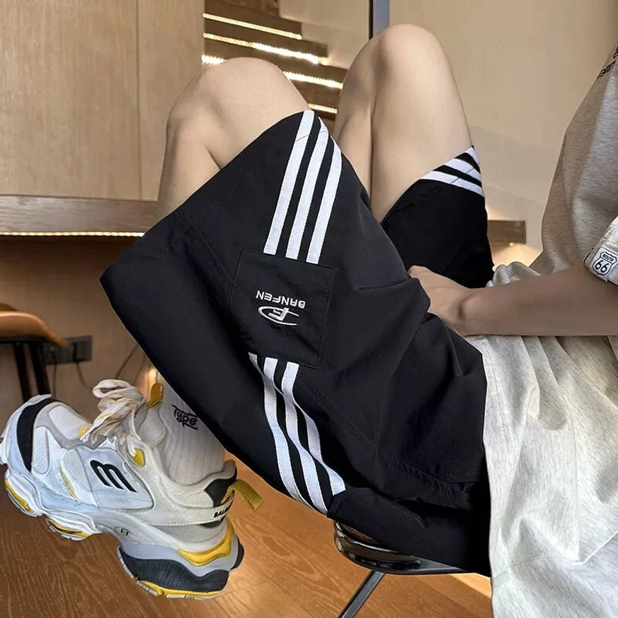 Loose-fit black streetwear tech shorts with three white side stripes, front patch detail, oversized tee, and chunky white sneakers