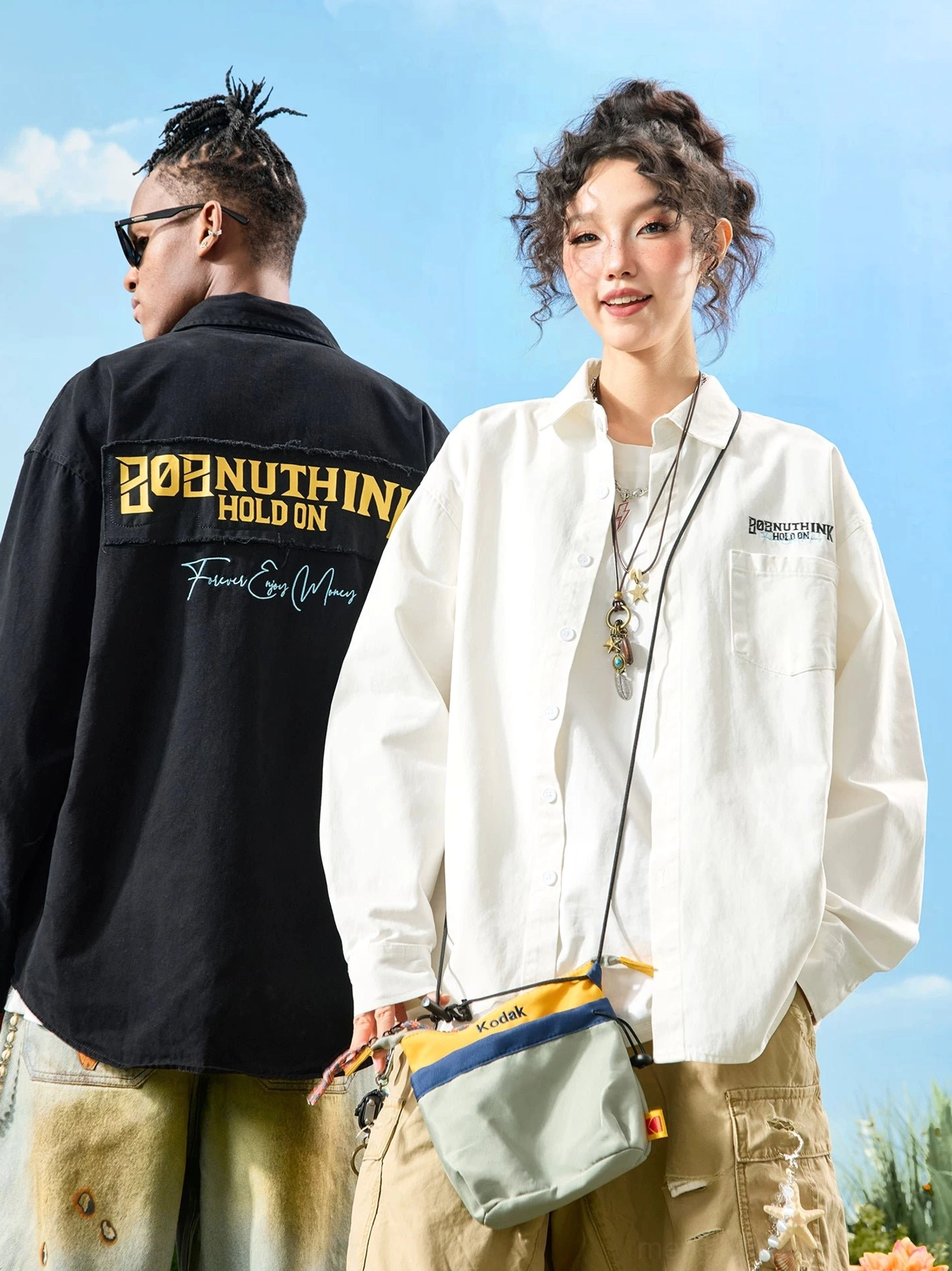 Two models wearing matching oversized embroidered button-up shirts with cargo pants, crossbody bag, and layered streetwear accessories