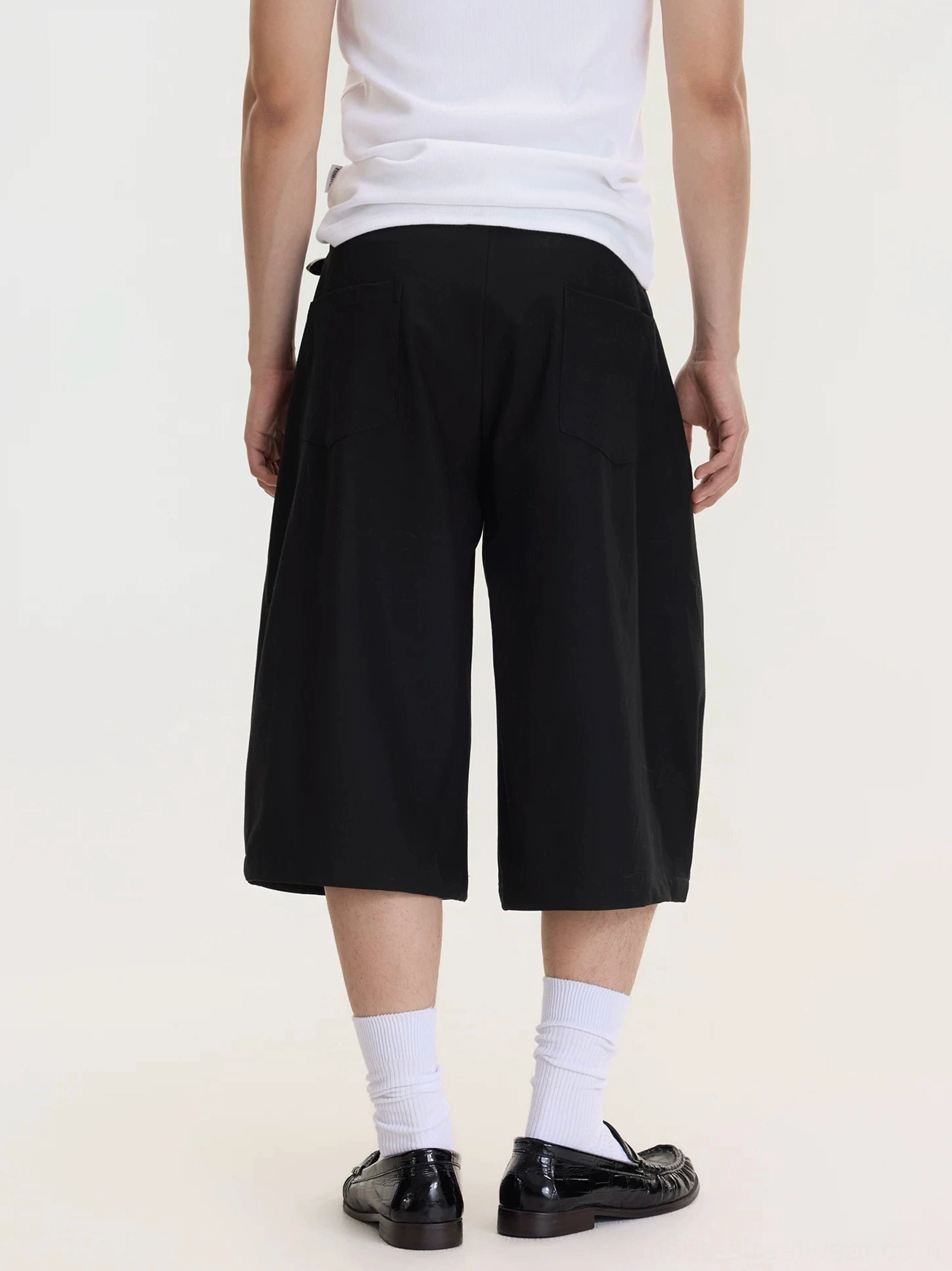 Back view of men's black cropped wide-leg culottes with clean matte fabric, rear patch pockets, and relaxed knee-length silhouette