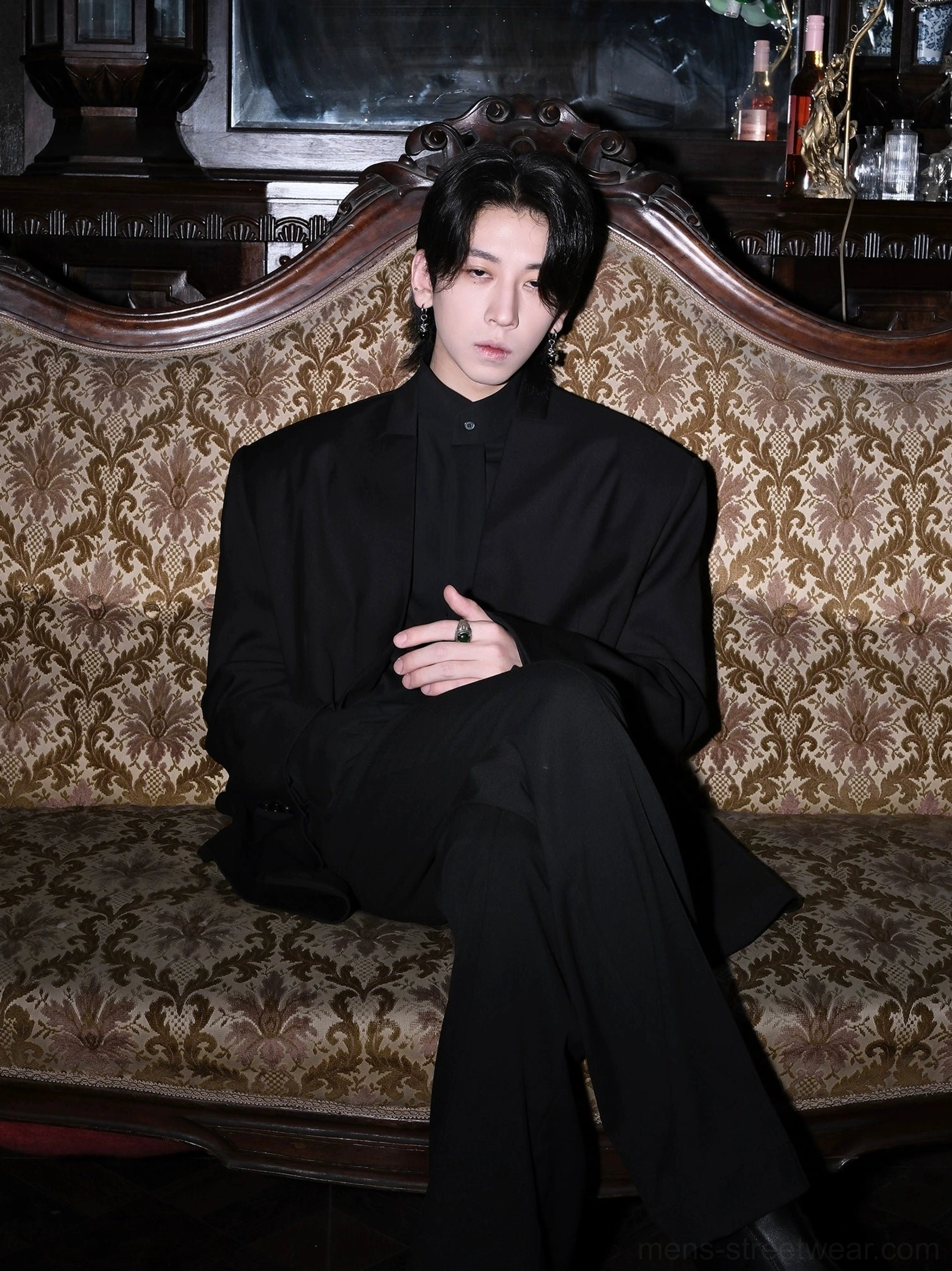 Men’s black minimalist collarless blazer shown in a seated editorial look inside a vintage room