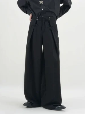 Men's black ultra wide-leg buckle pleated trousers shown from a low front angle with high waist, metal buckle details, deep front pleats, flap pockets, and oversized streetwear tailoring