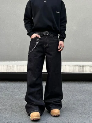 Front view of men's black wide-leg streetwear jeans styled with a black crewneck sweatshirt, leather belt, metal pocket chain, and urban sneakers against a concrete street wall