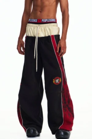 Black men's contrast panel wide-leg track trousers shown in a full front view with red side stripes, layered elastic waistband, drawstring closure, and streetwear silhouette