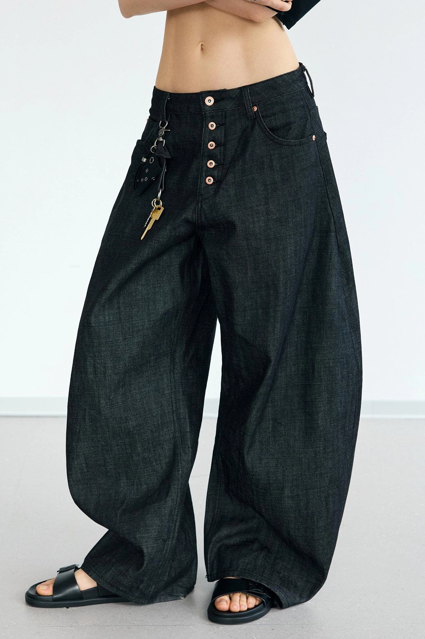 Low-angle detail view of men's curved wide-leg jeans showing the exposed button fly, gold-toned keychain hardware, and dark denim