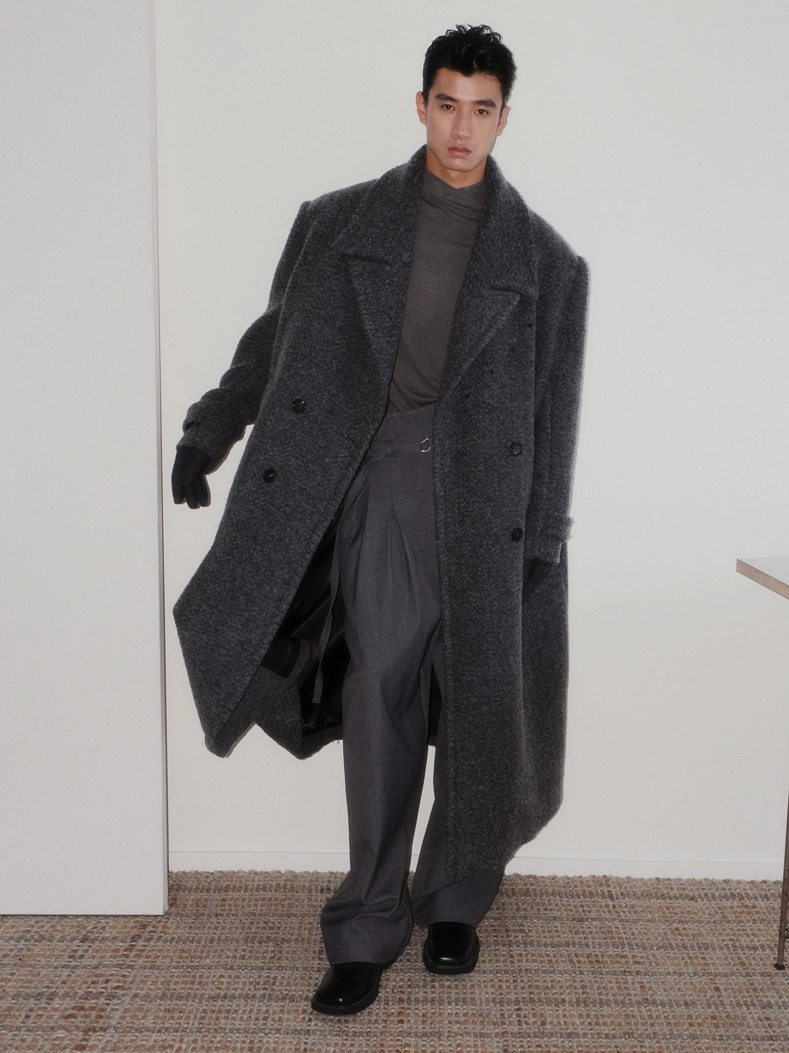 Side walking view of men's oversized dark gray bouclé wool long overcoat showing satin lining, double-breasted tailoring, and leather gloves
