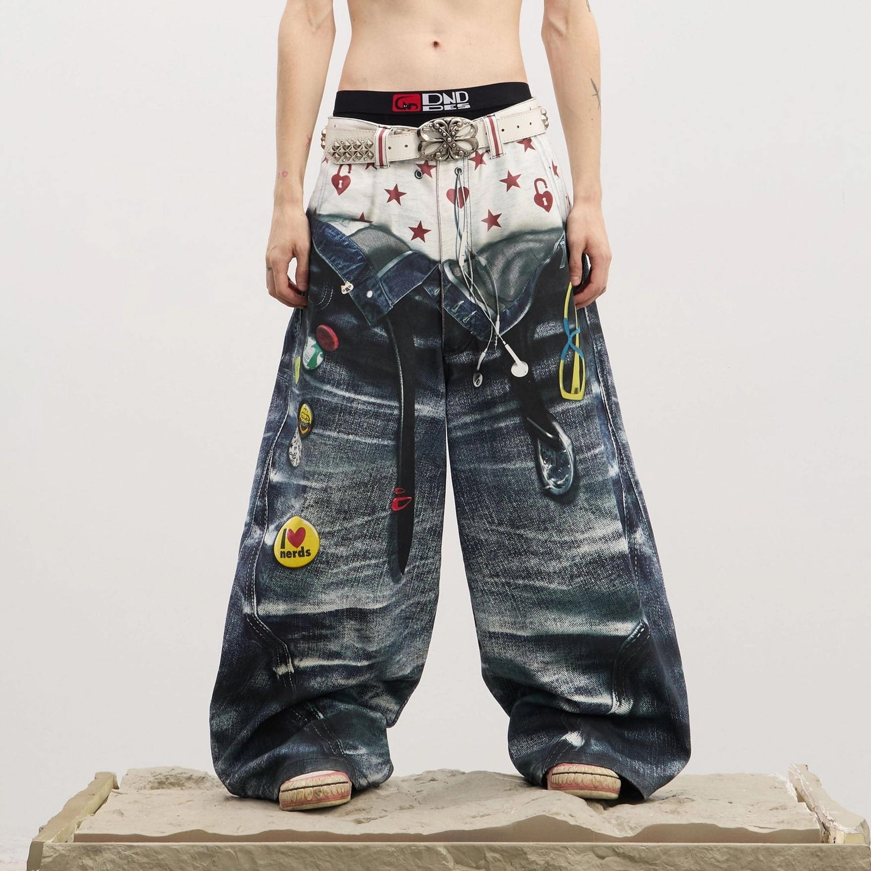 Front full-body view of men's dark wash sagging-illusion wide-leg jeans with printed boxer waistband, star and heart graphics, decorative belt, and enamel-style patches