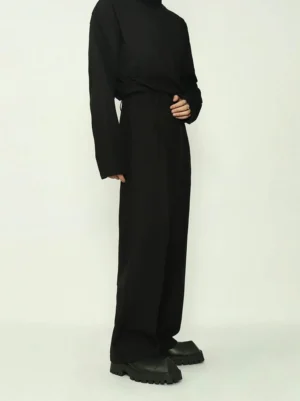 Men's high-waisted black wide straight-leg tailored trousers shown in left side full-body view with black turtleneck in white studio