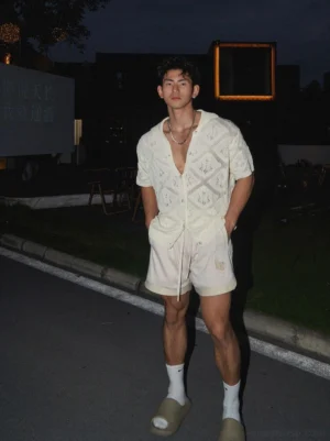 Full-body male model wearing a relaxed off-white crochet shirt with matching summer shorts white socks and neutral slides