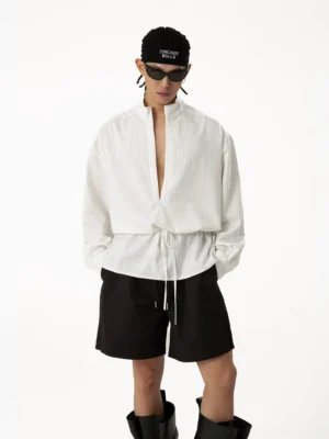 Front full-body view of men's off-white pinstripe plunge neck tunic shirt with stand collar, oversized fit, drawstring waist, black shorts, sunglasses, and chunky boots