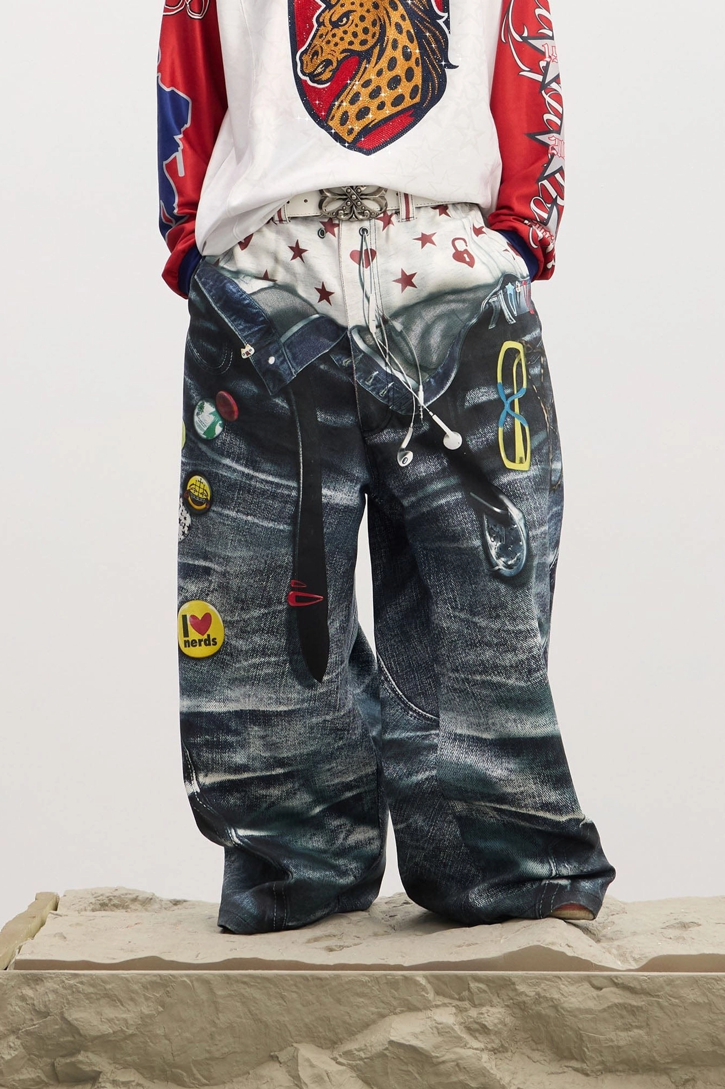 Front close-up of men's sagging-illusion wide-leg jeans showing printed star and heart waistband, enamel-style patches, denim texture, and ultra-baggy legs