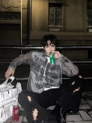Male model wearing a black sheer honeycomb mesh button-up shirt with printed typography, ripped black jeans, boots, and a graphic undershirt in a streetwear setting