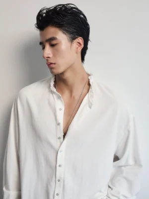 Close up of men's white ruffle band collar shirt with partial button placket, soft textured fabric, and silver necklace styling