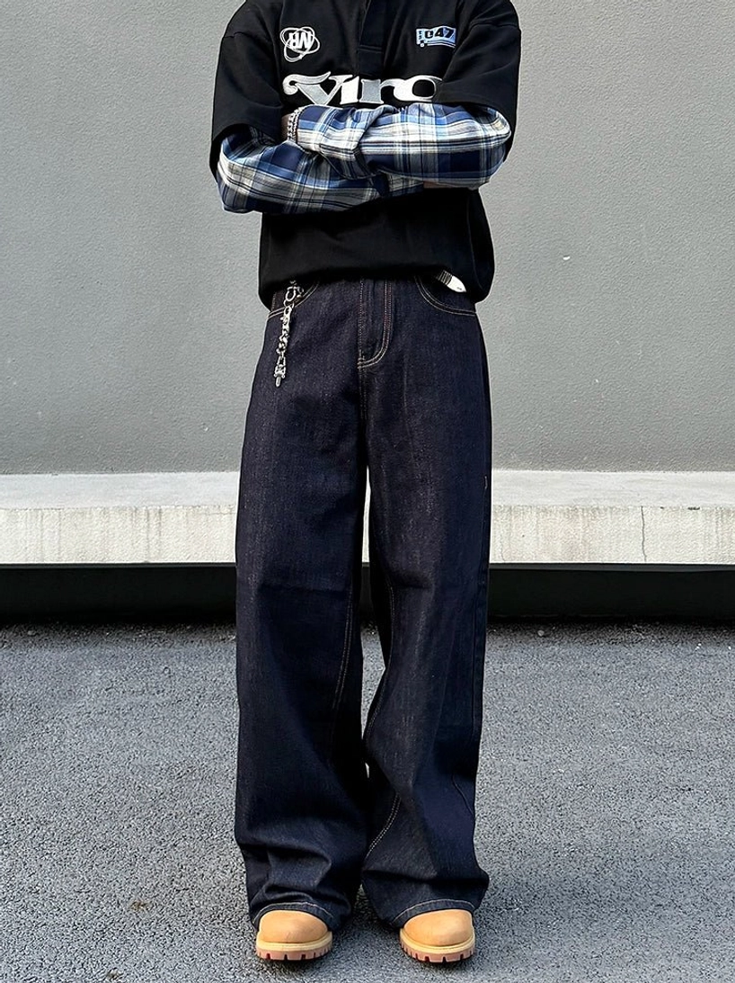 Men’s dark blue wide-leg stacked denim jeans styled with layered plaid shirt, graphic polo, waist chain, and work boots