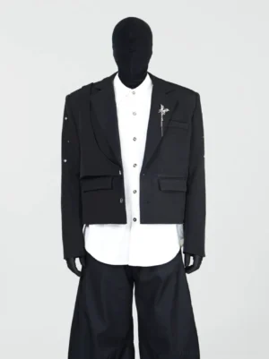 Front full-body view of a black layered cropped blazer with double-notched lapels, metallic lapel brooch, sleeve studs, and structured streetwear tailoring