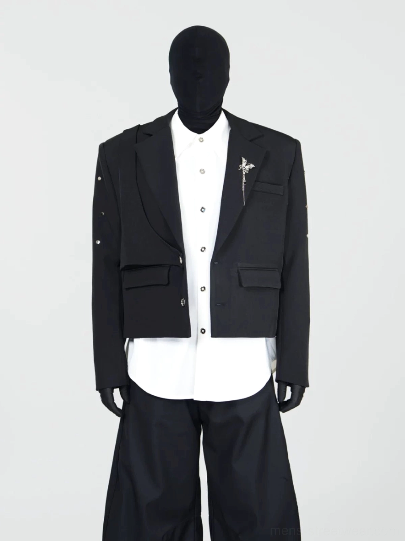 Front full-body view of a black layered cropped blazer with double-notched lapels, metallic lapel brooch, sleeve studs, and structured streetwear tailoring