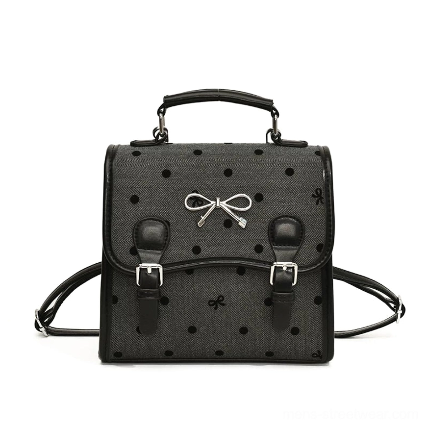 Front view of a dark grey polka dot bow satchel backpack with black faux leather trim, silver bow ornament, top handle, and convertible shoulder straps