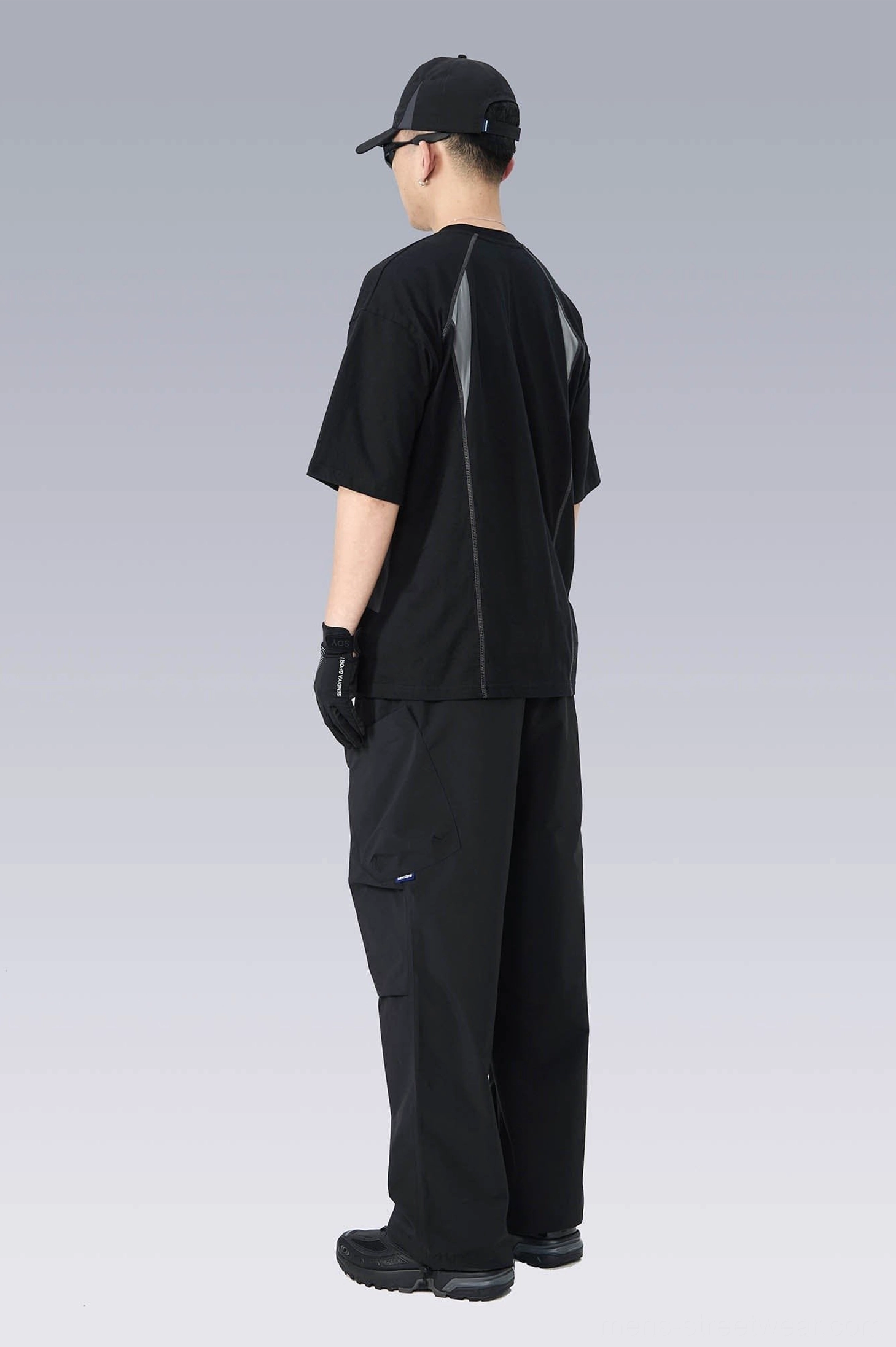 Rear side profile of a minimalist black techwear T-shirt with engineered seam lines, relaxed silhouette, and matching cargo pants