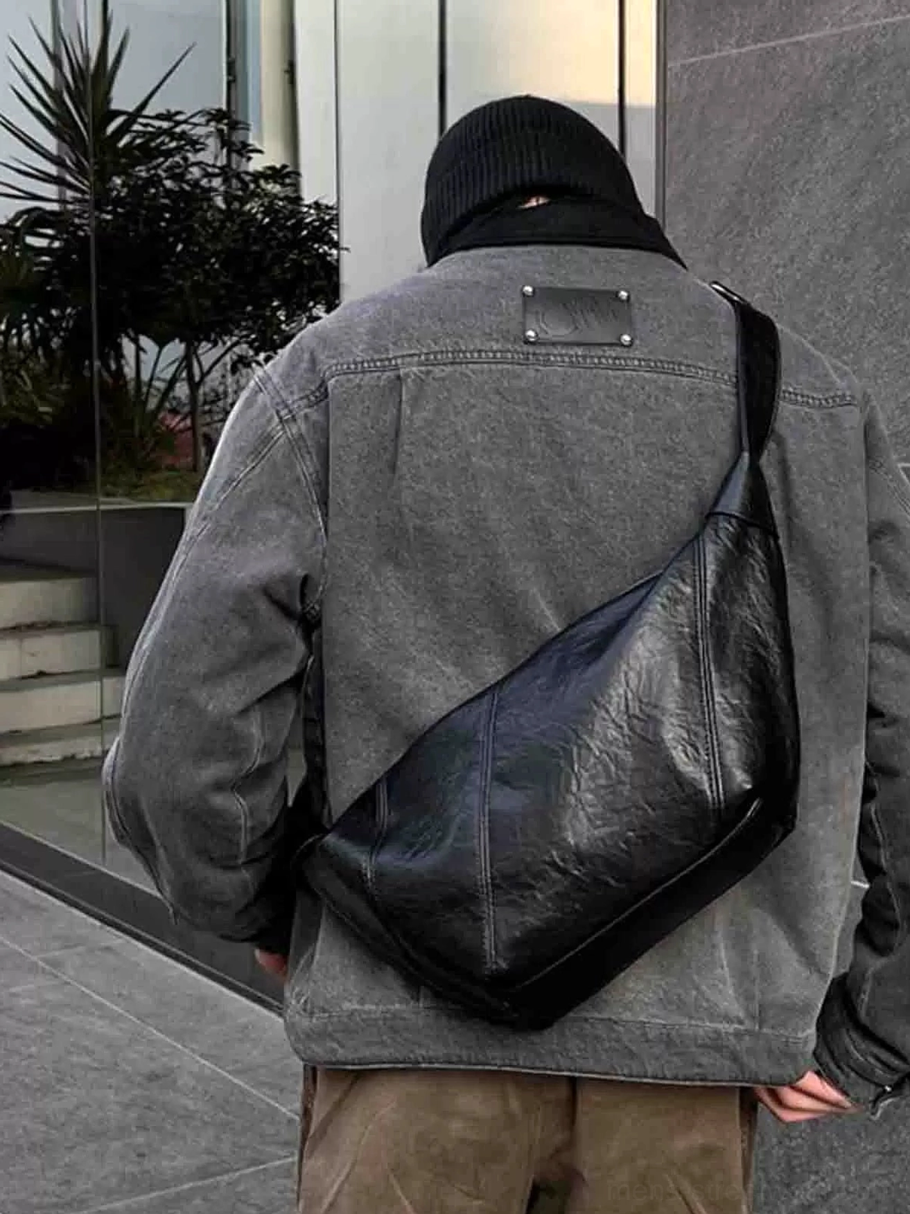 Black minimalist crossbody sling bag worn on the back with gray denim jacket and khaki pants in an urban plaza