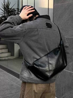 Black minimalist curved PU leather crossbody sling bag worn across the back with gray denim streetwear outfit outdoors